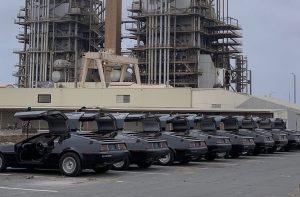 lineup of matte black DeLorean cars with gull-wing doors open, parked in front of an industrial power plant structure with pipes and metal frameworks in the background